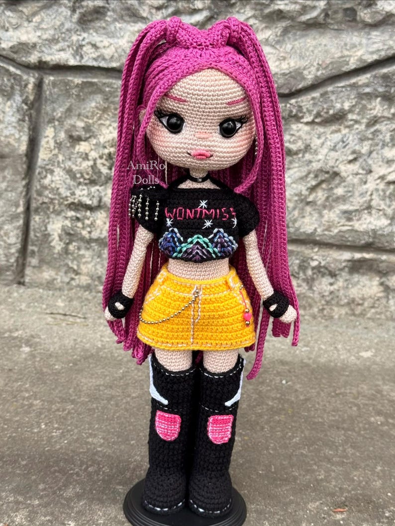 Puede incluir: Una mu&ntilde;eca de ganchillo hecha a mano con largo cabello rosa vibrante, que viste un top corto negro con el texto WONT MISS y un gr&aacute;fico de monta&ntilde;a, una minifalda amarilla brillante con una cadena plateada y botas negras altas con detalles.