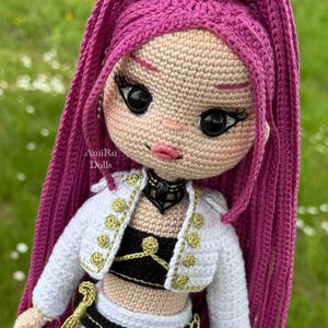 Puede incluir: Una mu&ntilde;eca tejida a mano con largo cabello magenta vibrante peinado en una coleta alta. La mu&ntilde;eca tiene grandes ojos negros, labios rosados y piel clara. Viste una chaqueta corta blanca con botones dorados, un top negro y una falda blanca.