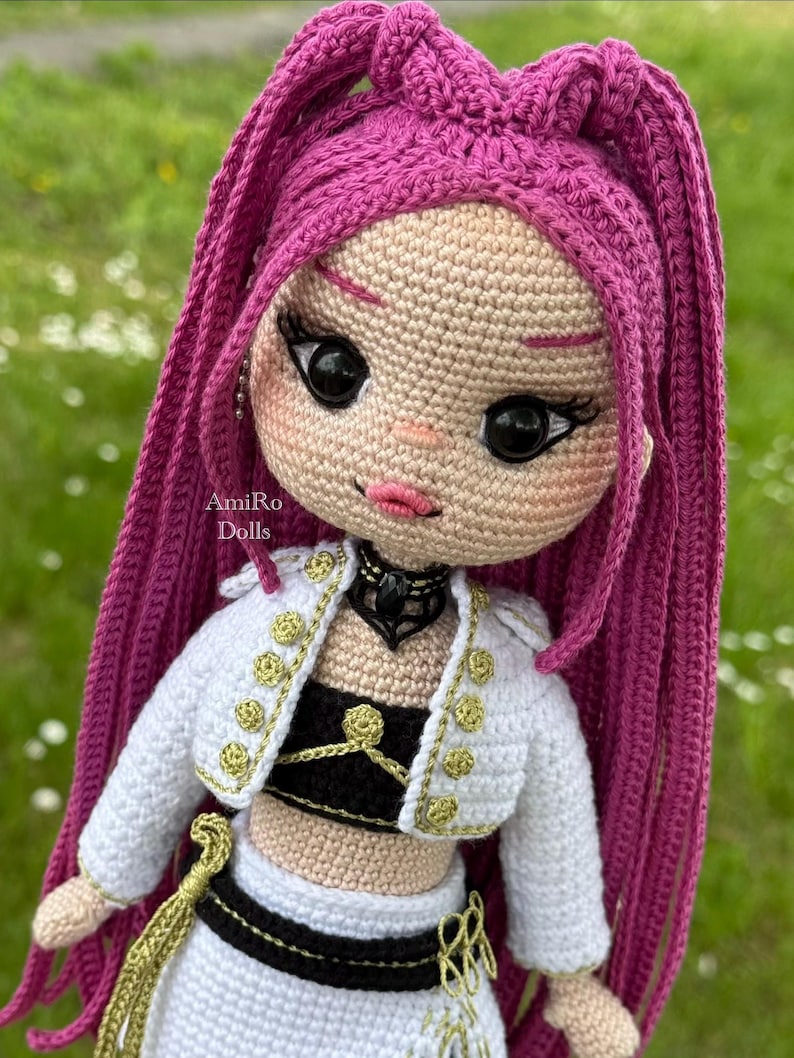 Puede incluir: Una mu&ntilde;eca tejida a mano con largo cabello magenta vibrante peinado en una coleta alta. La mu&ntilde;eca tiene grandes ojos negros, labios rosados y piel clara. Viste una chaqueta corta blanca con botones dorados, un top negro y una falda blanca.