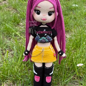 Puede incluir: Una mu&ntilde;eca tejida a mano con largo cabello rosa vibrante, que viste un top corto negro con el texto WONTMISS, una minifalda amarilla con una cadena plateada y botas negras hasta la rodilla con detalles en rosa y blanco. Ojos negros grandes.
