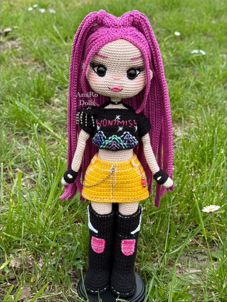 Puede incluir: Una mu&ntilde;eca tejida a mano con largo cabello rosa vibrante, que viste un top corto negro con el texto WONTMISS, una minifalda amarilla con una cadena plateada y botas negras hasta la rodilla con detalles en rosa y blanco. Ojos negros grandes.