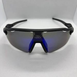 Op de afbeelding: Zwarte sportzonnebril met spiegelglazen en blauwe accenten. De zonnebril heeft een strak, modern design met een gebogen montuur en de tekst "Vista Market".