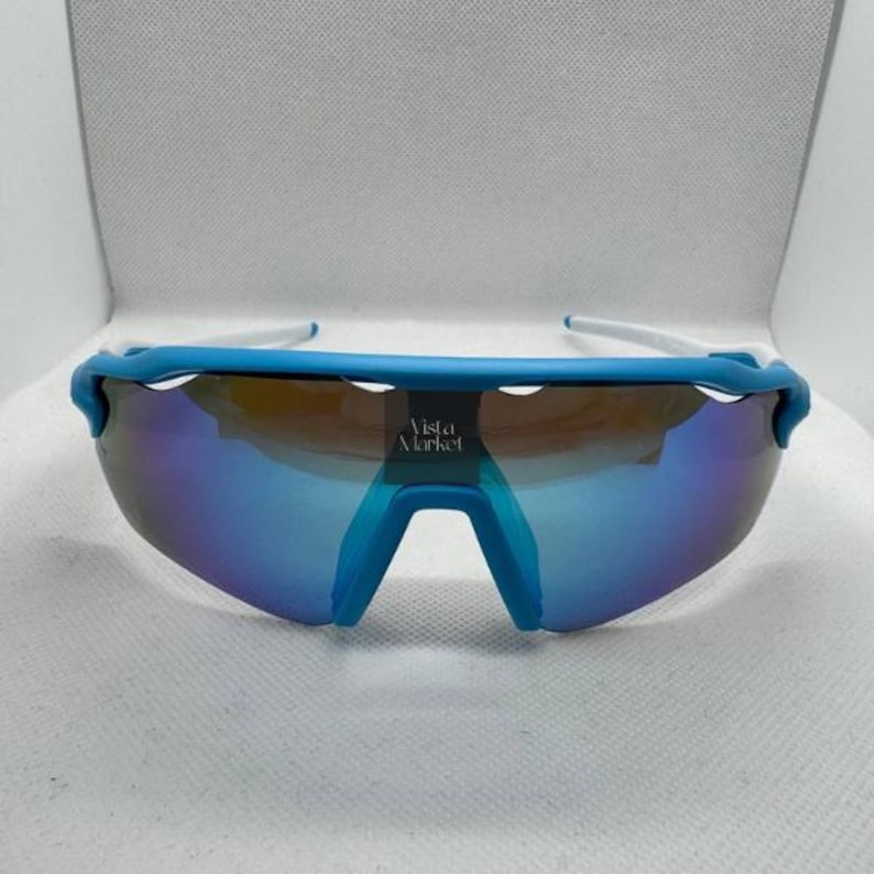 Op de afbeelding: Sport zonnebril met een blauw montuur en spiegelglazen. De zonnebril heeft een witte poot en een blauwe brug. De glazen reflecteren een kleurengradient. De tekst "Vista Market" is zichtbaar op de lens.