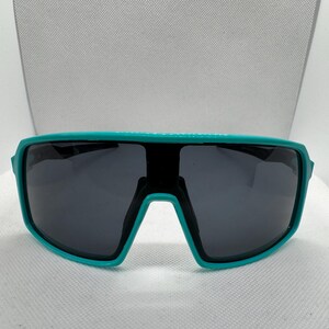 Puede incluir: Gafas de sol con montura turquesa y lentes gris oscuro. Las gafas tienen un dise&ntilde;o deportivo envolvente. La montura es de un color turquesa vibrante y las lentes son de color gris oscuro, lo que proporciona un aspecto elegante y moderno. Adecuadas para actividades al aire libre.