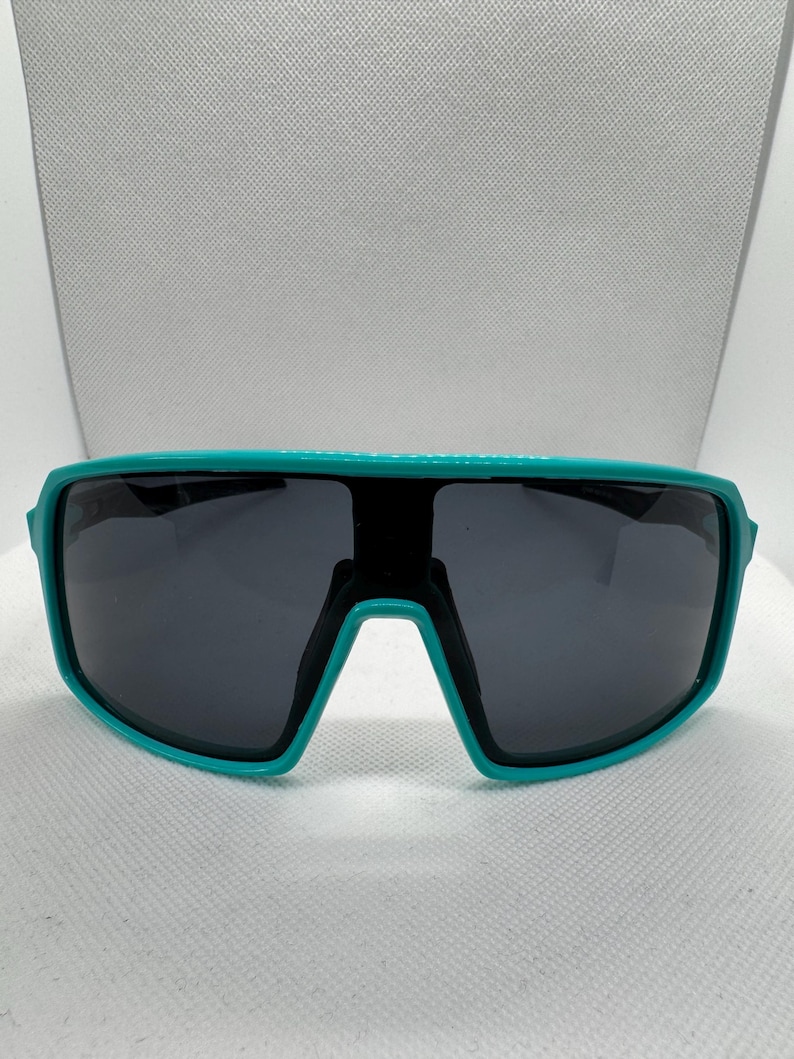 Puede incluir: Gafas de sol con montura turquesa y lentes gris oscuro. Las gafas tienen un dise&ntilde;o deportivo envolvente. La montura es de un color turquesa vibrante y las lentes son de color gris oscuro, lo que proporciona un aspecto elegante y moderno. Adecuadas para actividades al aire libre.