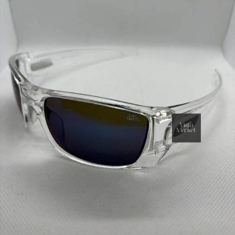 May include: Clear-framed sunglasses with dark blue lenses. The sunglasses have a rectangular shape and a sleek, modern design. The text "Vista Market" is visible on the right temple.