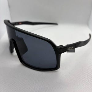 Puede incluir: Gafas de sol negras envolventes con lentes grises oscuros y montura negra mate. Las gafas de sol tienen un dise&ntilde;o moderno. El texto "Vista Market" est&aacute; impreso en la patilla.