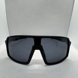 Puede incluir: Gafas de sol negras con una montura gruesa y rectangular y lentes de color gris oscuro. Las gafas de sol tienen un dise&ntilde;o moderno y deportivo, adecuado para actividades al aire libre. Las lentes son anchas, ofreciendo una amplia cobertura.