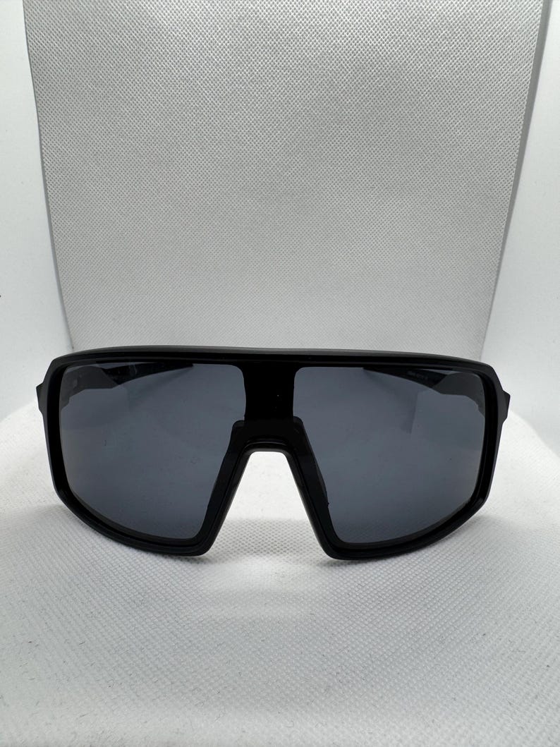 Puede incluir: Gafas de sol negras con una montura gruesa y rectangular y lentes de color gris oscuro. Las gafas de sol tienen un dise&ntilde;o moderno y deportivo, adecuado para actividades al aire libre. Las lentes son anchas, ofreciendo una amplia cobertura.
