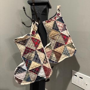 May include: Two quilted Christmas stockings with patchwork patterns in red, blue, green, and tan. Each stocking has a loop with a red button and a white, fluffy trim. They hang from a black, decorative post with hooks.