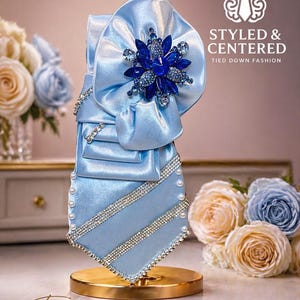 May include: A light blue satin tie with a large bow and a dark blue jeweled flower. The tie is decorated with rhinestones and pearls. The text "STYLED & CENTERED TIED DOWN FASHION" is visible. The text "Text 1-843-900-1072" is also visible.