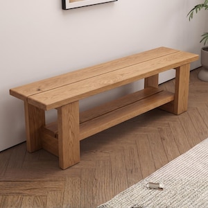 May include: A rectangular wooden bench featuring a natural light brown wood grain finish. The bench is constructed with four thick, sturdy legs and includes a lower shelf positioned between the legs, providing additional storage space beneath the seat.