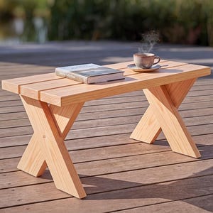 May include: A light-colored wooden bench featuring a slatted top and two crossed-leg supports at each end. The bench is constructed from natural wood planks and displays a clear grain pattern throughout the entire structure of the furniture piece.