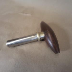 Puede incluir: Un tapón de botella de vino con un mango de madera redondeado de color marrón oscuro. El tapón tiene un vástago de metal plateado y un anillo de color latón donde el mango se une al vástago. El tapón está diseñado para encajar en una botella de vino.
