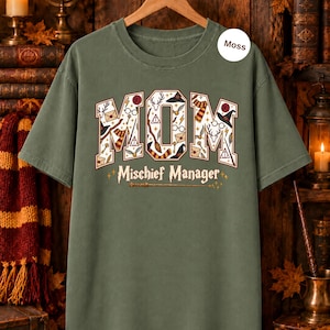 Op de afbeelding: Een mosgroen T-shirt met korte mouwen en het woord MOM in grote blokletters, gevuld met diverse magische iconen zoals hoeden, brillen en toverstokken. Onder het woord MOM staat de tekst Mischief Manager met een afbeelding van een toverstok.