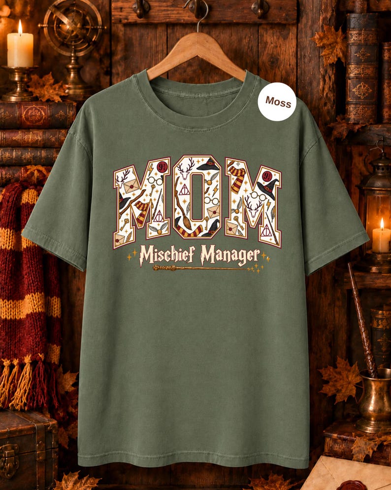 Op de afbeelding: Een mosgroen T-shirt met korte mouwen en het woord MOM in grote blokletters, gevuld met diverse magische iconen zoals hoeden, brillen en toverstokken. Onder het woord MOM staat de tekst Mischief Manager met een afbeelding van een toverstok.