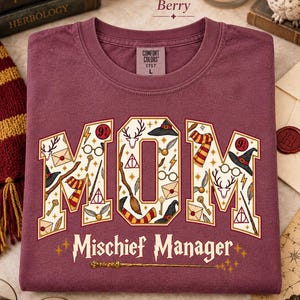 Op de afbeelding: Een bessenkleurig T-shirt met ronde hals en een Comfort Colors label in maat L. De voorkant toont het woord MOM in grote blokletters gevuld met magische iconen zoals toverstokken, hoeden en brillen, met de tekst Mischief Manager eronder.
