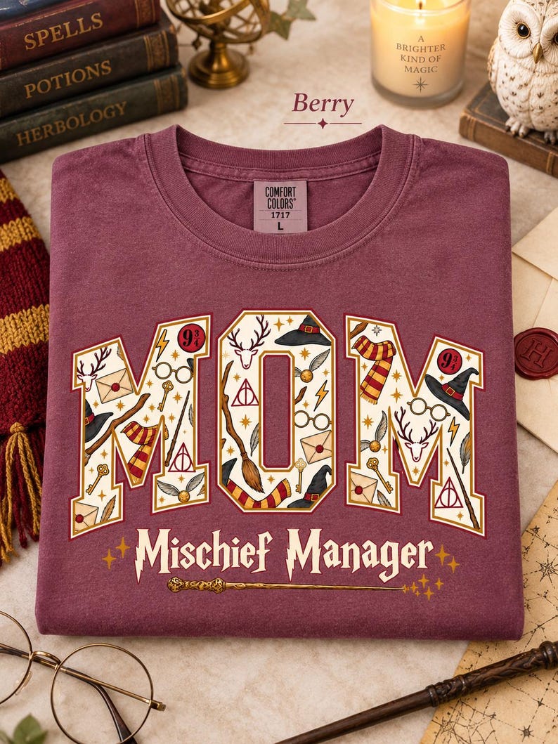 Op de afbeelding: Een bessenkleurig T-shirt met ronde hals en een Comfort Colors label in maat L. De voorkant toont het woord MOM in grote blokletters gevuld met magische iconen zoals toverstokken, hoeden en brillen, met de tekst Mischief Manager eronder.