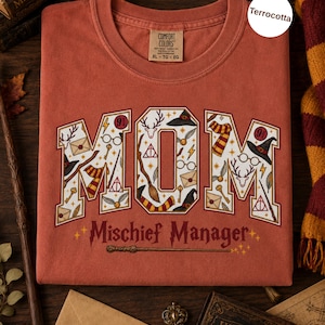 Op de afbeelding: Een terracotta-kleurig t-shirt met het woord MOM in grote blokletters gevuld met magische iconen zoals toverstokken, brillen en sjaals. Onder het woord MOM staat de tekst Mischief Manager in een donkerrode kleur met een toverstokafbeelding.