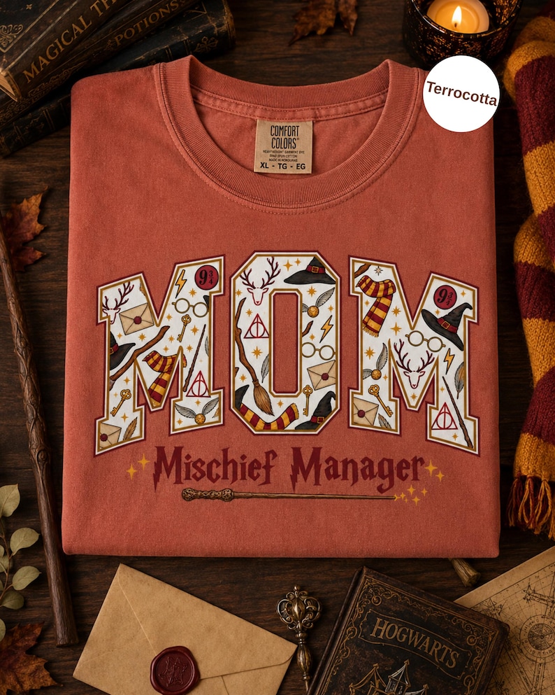 Op de afbeelding: Een terracotta-kleurig t-shirt met het woord MOM in grote blokletters gevuld met magische iconen zoals toverstokken, brillen en sjaals. Onder het woord MOM staat de tekst Mischief Manager in een donkerrode kleur met een toverstokafbeelding.