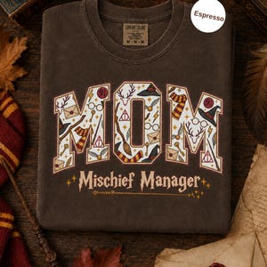 Op de afbeelding: Een espresso-kleurig t-shirt met het woord MOM in grote blokletters gevuld met magische iconen zoals toverstokken, hoeden en brillen. Onder het woord MOM staat de tekst Mischief Manager in een gestileerd lettertype met een toverstokafbeelding.