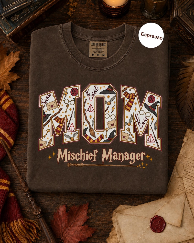 Op de afbeelding: Een espresso-kleurig t-shirt met het woord MOM in grote blokletters gevuld met magische iconen zoals toverstokken, hoeden en brillen. Onder het woord MOM staat de tekst Mischief Manager in een gestileerd lettertype met een toverstokafbeelding.