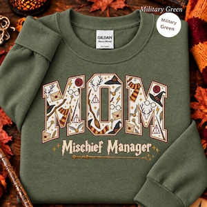 Op de afbeelding: Een militair groen sweatshirt met ronde hals en het woord MOM in grote blokletters gevuld met diverse magische iconen zoals toverstokken, brillen en sjaals. Onder het woord MOM staat de tekst Mischief Manager in een sierlijk lettertype.