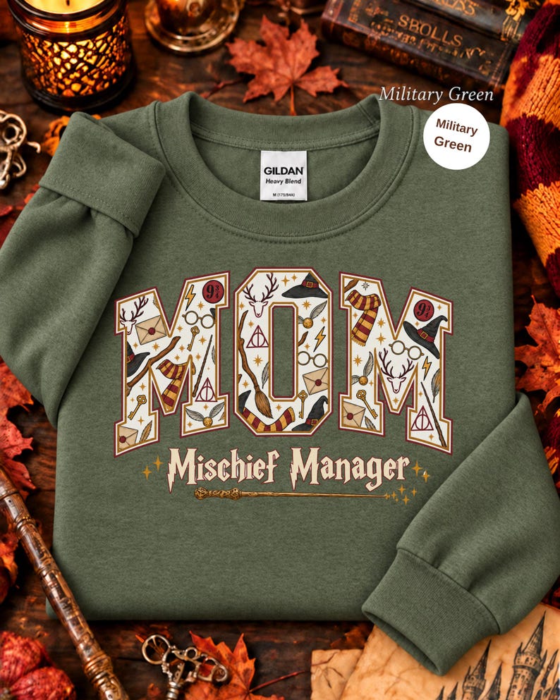 Op de afbeelding: Een militair groen sweatshirt met ronde hals en het woord MOM in grote blokletters gevuld met diverse magische iconen zoals toverstokken, brillen en sjaals. Onder het woord MOM staat de tekst Mischief Manager in een sierlijk lettertype.