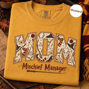 Op de afbeelding: Een mosterdgeel t-shirt met het woord MOM in grote blokletters gevuld met diverse magische iconen zoals toverstokken, brillen en hoeden. Onder het woord MOM staat de tekst Mischief Manager in een donkerbruin lettertype.