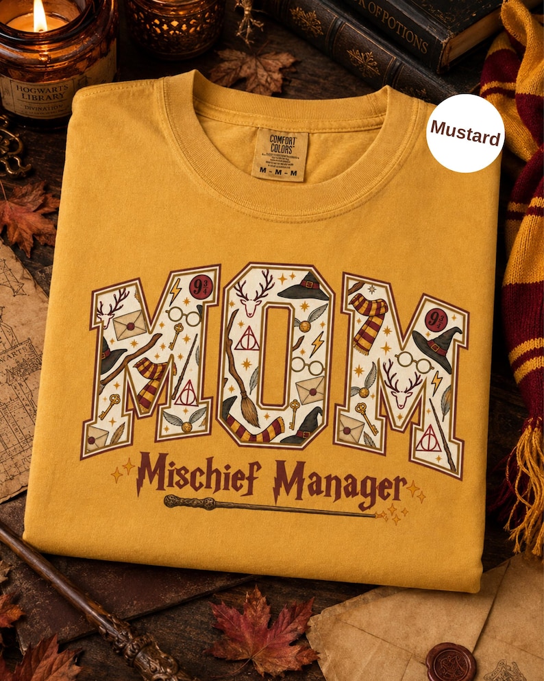 Op de afbeelding: Een mosterdgeel t-shirt met het woord MOM in grote blokletters gevuld met diverse magische iconen zoals toverstokken, brillen en hoeden. Onder het woord MOM staat de tekst Mischief Manager in een donkerbruin lettertype.