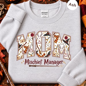 Op de afbeelding: Een asgrijze sweater met ronde hals waarop het woord MOM in grote blokletters staat, gevuld met diverse magische iconen zoals toverstokken, brillen en sjaals. Onder het woord MOM staat de tekst Mischief Manager in een donkerrode kleur.