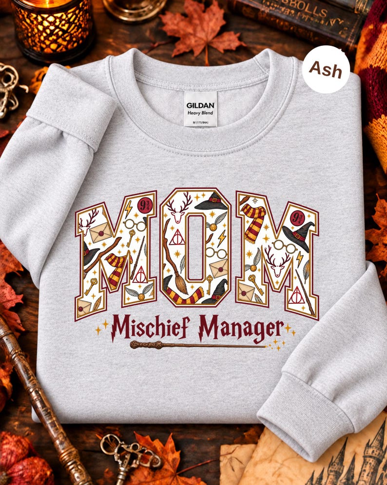 Op de afbeelding: Een asgrijze sweater met ronde hals waarop het woord MOM in grote blokletters staat, gevuld met diverse magische iconen zoals toverstokken, brillen en sjaals. Onder het woord MOM staat de tekst Mischief Manager in een donkerrode kleur.
