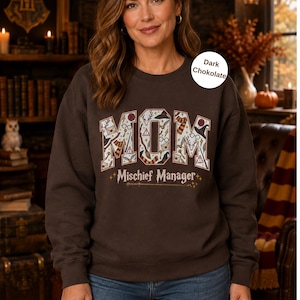 Op de afbeelding: Een donkerbruin sweatshirt met ronde hals met het woord MOM in grote letters gevuld met diverse magische iconen en de tekst Mischief Manager eronder. Het sweatshirt heeft lange mouwen en een geribbelde kraag, boorden en zoom.