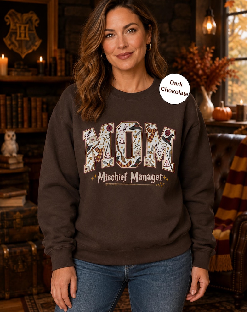 Op de afbeelding: Een donkerbruin sweatshirt met ronde hals met het woord MOM in grote letters gevuld met diverse magische iconen en de tekst Mischief Manager eronder. Het sweatshirt heeft lange mouwen en een geribbelde kraag, boorden en zoom.