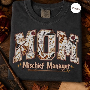 Op de afbeelding: Een antracietgrijs t-shirt met het woord MOM in grote blokletters gevuld met magische iconen zoals toverstokken, hoeden en brillen. Onder het woord MOM staat de tekst Mischief Manager in een gestileerd lettertype met een toverstokafbeelding.