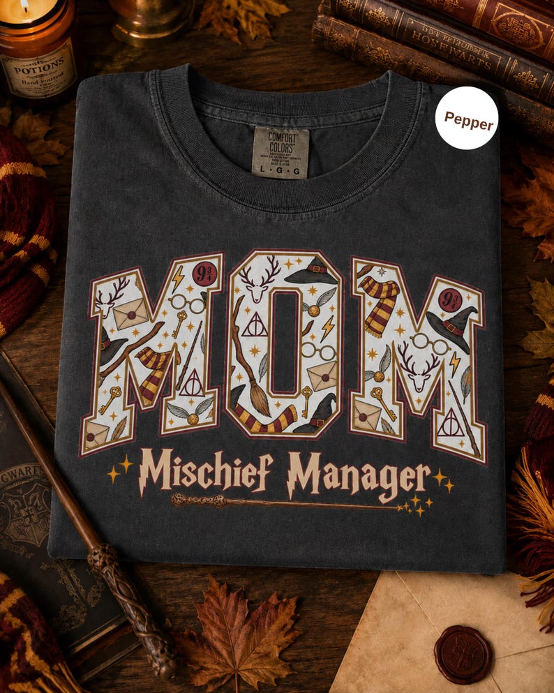 Op de afbeelding: Een antracietgrijs t-shirt met het woord MOM in grote blokletters gevuld met magische iconen zoals toverstokken, hoeden en brillen. Onder het woord MOM staat de tekst Mischief Manager in een gestileerd lettertype met een toverstokafbeelding.