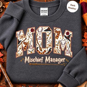 Op de afbeelding: Een donkergrijs gem&ecirc;leerd sweatshirt met ronde hals en het woord MOM in grote letters gevuld met magische iconen zoals toverstokken, hoeden en brillen. Onder het woord MOM staat de tekst Mischief Manager met een toverstokafbeelding.