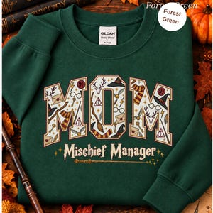 Op de afbeelding: Een bosgroene sweater met ronde hals en het woord MOM in grote blokletters gevuld met diverse magische iconen zoals toverstokken, brillen en hoeden. Onder het woord MOM staat de tekst Mischief Manager in een gestileerd lettertype.