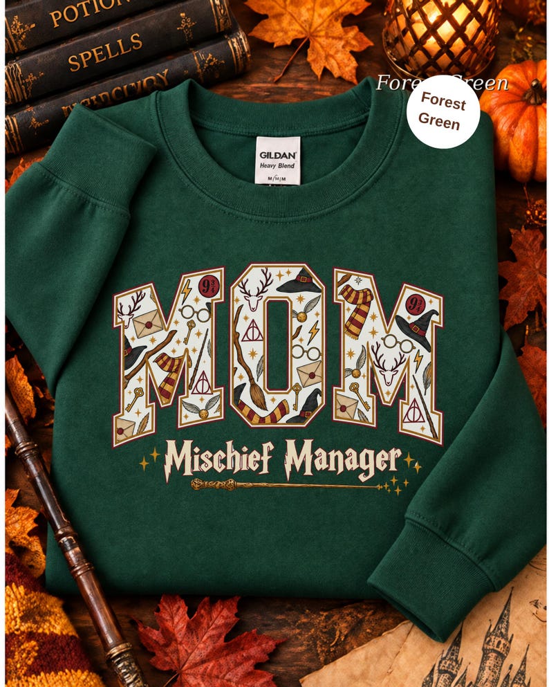 Op de afbeelding: Een bosgroene sweater met ronde hals en het woord MOM in grote blokletters gevuld met diverse magische iconen zoals toverstokken, brillen en hoeden. Onder het woord MOM staat de tekst Mischief Manager in een gestileerd lettertype.