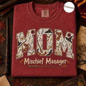 Op de afbeelding: Een karmozijnrood T-shirt met ronde hals en het woord MOM in grote blokletters, gevuld met magische iconen zoals een hert, een bril, een toverstaf en een bezem. Daaronder staat de tekst Mischief Manager in een gestileerd lettertype.
