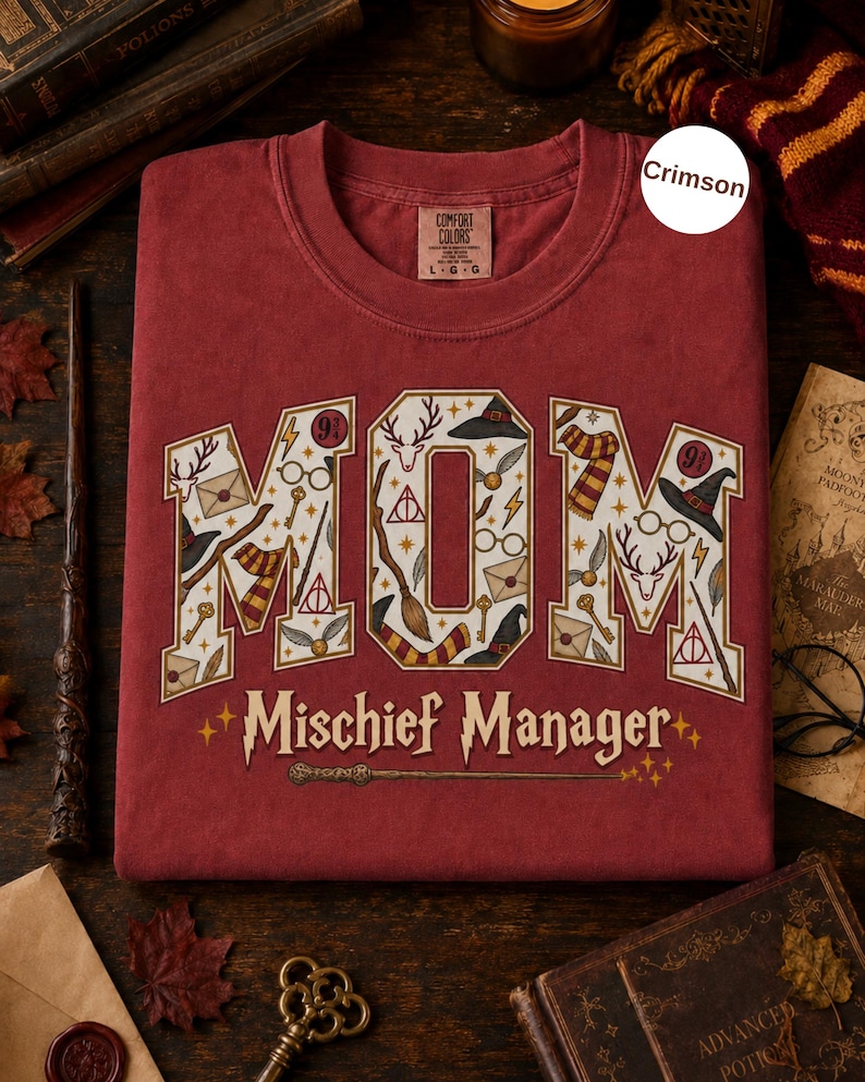 Op de afbeelding: Een karmozijnrood T-shirt met ronde hals en het woord MOM in grote blokletters, gevuld met magische iconen zoals een hert, een bril, een toverstaf en een bezem. Daaronder staat de tekst Mischief Manager in een gestileerd lettertype.