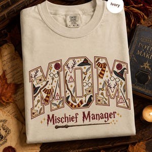 Op de afbeelding: Een ivoorkleurig T-shirt met korte mouwen en het woord MOM in grote blokletters gevuld met tovenaarsillustraties zoals brillen, toverstokken, hoeden en sjaals. Onder het woord MOM staat de tekst Mischief Manager in een gestileerd lettertype.