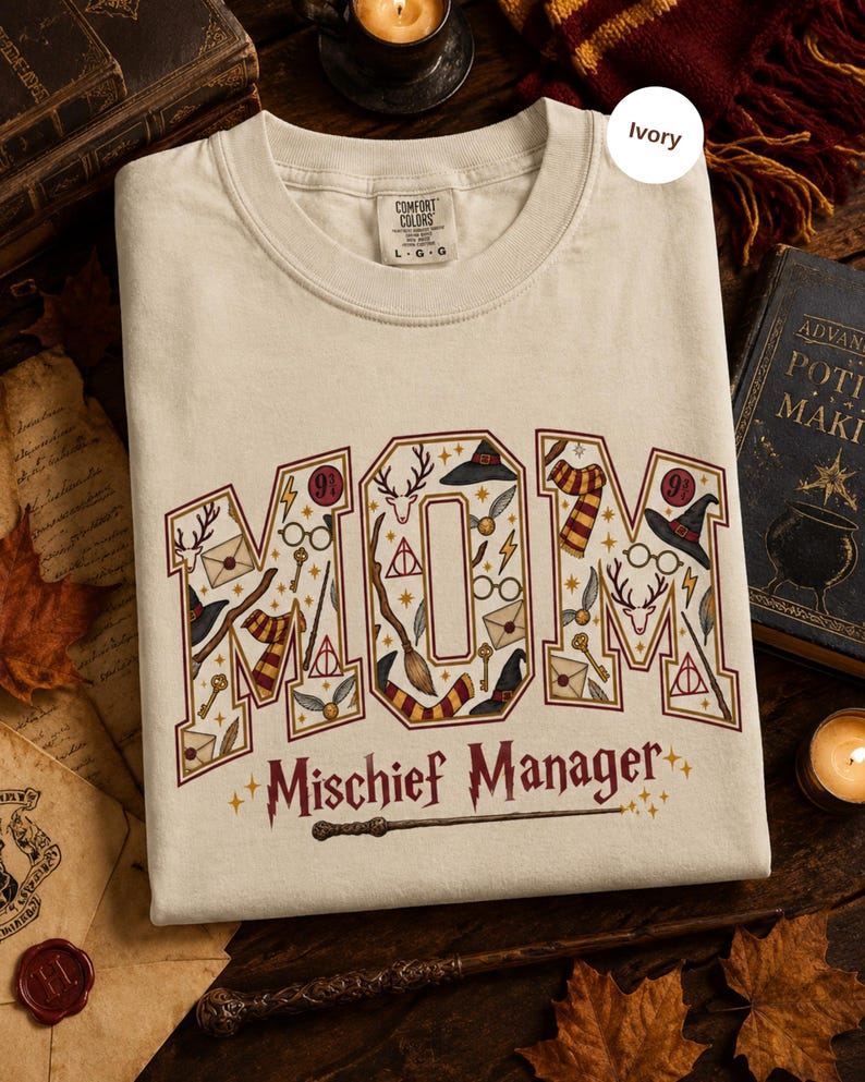Op de afbeelding: Een ivoorkleurig T-shirt met korte mouwen en het woord MOM in grote blokletters gevuld met tovenaarsillustraties zoals brillen, toverstokken, hoeden en sjaals. Onder het woord MOM staat de tekst Mischief Manager in een gestileerd lettertype.