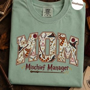 Op de afbeelding: Een zeegroen t-shirt met het woord MOM in grote blokletters gevuld met diverse magische iconen, waaronder toverstokken, brillen, hoeden en enveloppen. Onder het woord MOM staat de tekst Mischief Manager gedrukt in een donkerrode kleur.