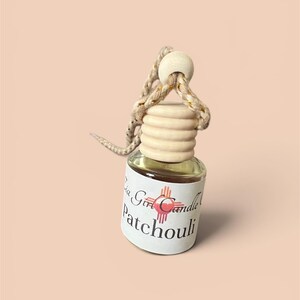 May include: A small glass bottle with a wooden cap and a woven rope for hanging. The bottle contains a clear liquid and a label that reads "La Girl Candle Patchouli". The background is a solid peach color.