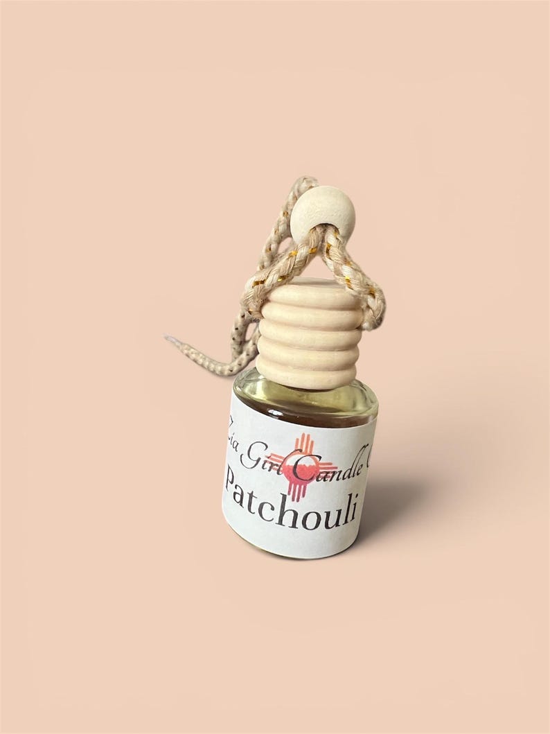 K&ouml;nnte beinhalten: Kleine Glasflasche mit Holzverschluss und geflochtener Schnur zum Aufh&auml;ngen. Enth&auml;lt eine klare Fl&uuml;ssigkeit und ein Etikett mit der Aufschrift "La Girl Candle Patchouli". Der Hintergrund ist einfarbig pfirsichfarben.