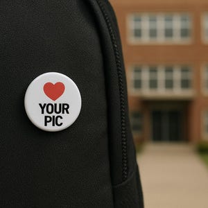 May include: A white button with a red heart and the text "YOUR PIC" is pinned to a black backpack. The button is round and has a glossy finish. The background is a blurred view of a brick building.