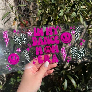 May include: A clear, rectangular sign with the text "IN MY DANCE MOM ERA" in bright pink, along with smiley faces, lightning bolts, stars, and checkered bows. The sign is held in front of a green leafy background.