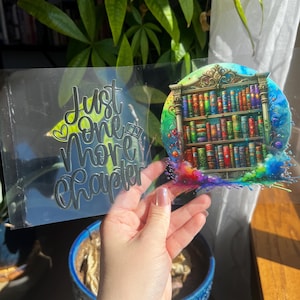 May include: A clear bookmark with the text "Just One More Chapter" in silver lettering. The bookmark features a colorful illustration of a bookshelf filled with books. The design includes a rainbow splash effect.