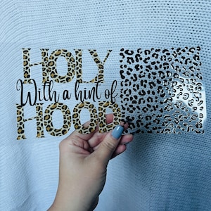 May include: A clear rectangular sticker with the text "HOLY With a hint of HOOD" in black and leopard print. The right side of the sticker has a leopard print design. The sticker is held up by a hand.